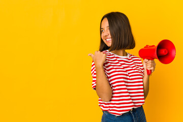 Young hispanic woman holding a megaphone points with thumb finger away, laughing and carefree.