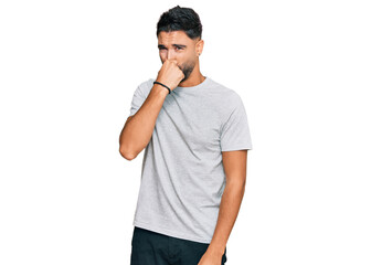 Young man with beard wearing casual grey tshirt smelling something stinky and disgusting, intolerable smell, holding breath with fingers on nose. bad smell
