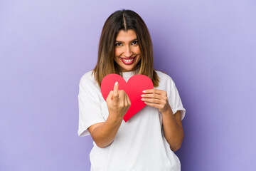 Young indian woman holding a valentines day heart isolated pointing with finger at you as if inviting come closer.
