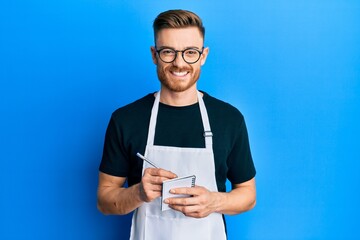 Young redhead man wearing waiter apron taking order smiling with a happy and cool smile on face. showing teeth. - obrazy, fototapety, plakaty