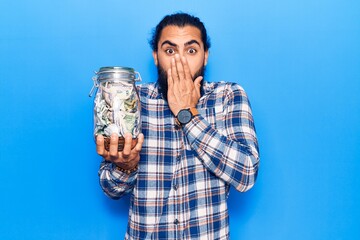 Young arab man holding jar with savings covering mouth with hand, shocked and afraid for mistake....