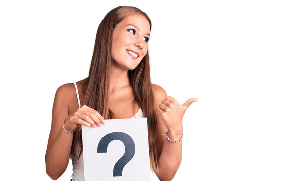 Young beautiful hispanic woman holding question mark pointing thumb up to the side smiling happy with open mouth