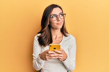 Young brunette woman using smartphone over yellow background smiling looking to the side and...