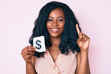 Beautiful african woman holding dollar symbol paper smiling with an idea or question pointing finger with happy face, number one