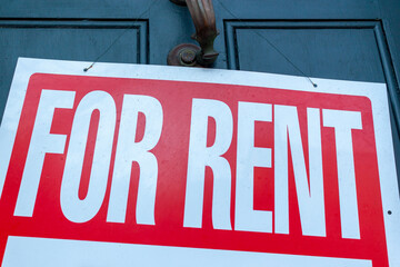 A large For Rent sign (white text on red background) is attached to  metal door handle on the front door of an old townhome. A versatile image for real estate, housing market, economy related themes.