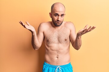 Young handsome bald man wearing swimwear shirtless clueless and confused with open arms, no idea and doubtful face.