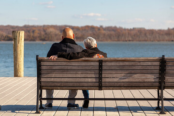 an elderly caucasian woman with short gray hair and a younger African American man with shaved head are sitting on a bench on a pier looking at sea. Image for interracial couple with age difference.