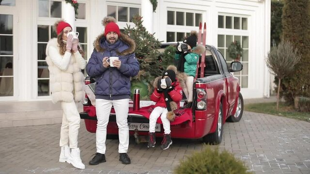 Stock video of stylish parents and two trendy kids sitting on back of red car drinking hot tea or coffee together outdoors. Drinking hot beverage from mugs to warm up in winter.