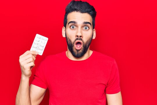 Young handsome man with beard holding pills scared and amazed with open mouth for surprise, disbelief face