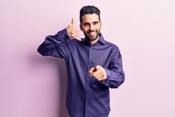 Young handsome man with beard wearing casual shirt smiling doing talking on the telephone gesture and pointing to you. call me.