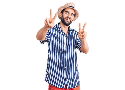 Young handsome man with beard wearing summer hat and striped shirt smiling looking to the camera showing fingers doing victory sign. number two.