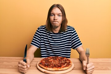 Handsome caucasian man with long hair eating tasty pepperoni pizza skeptic and nervous, frowning upset because of problem. negative person.