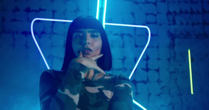 Skilled model in trendy green long sleeve shirt dances techno professionally against modern bright blue neon background closeup