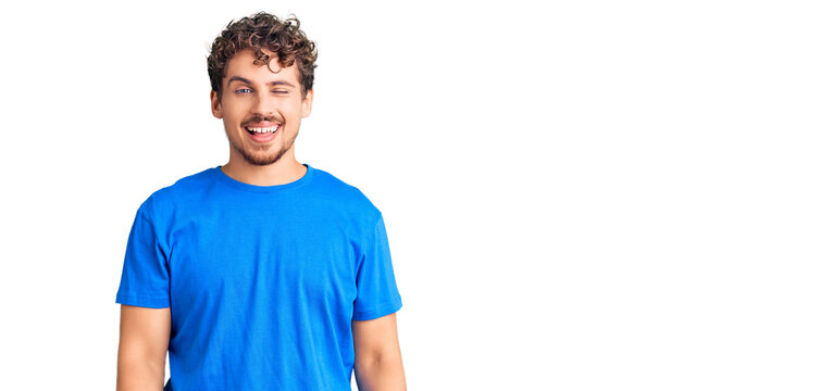 Young handsome man with curly hair wearing casual clothes winking looking at the camera with sexy expression, cheerful and happy face.