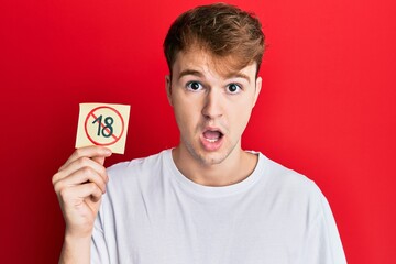 Young caucasian man holding under 18 prohibition sticker scared and amazed with open mouth for surprise, disbelief face