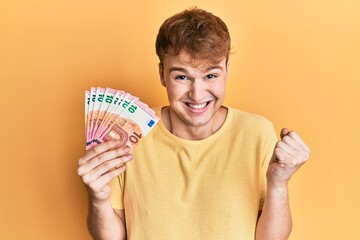 Young caucasian man holding bunch of 10 euro banknotes screaming proud, celebrating victory and success very excited with raised arm