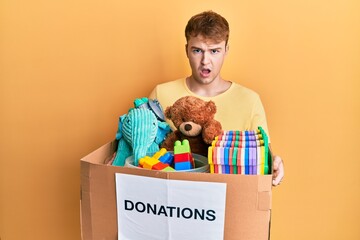 Young caucasian man holding donation box with toys in shock face, looking skeptical and sarcastic,...