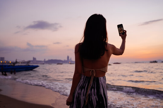 Young woman taking a selfie photo in sunset tropical beach sea view background with her mobile phone. Beautiful Asian girl photographing herself with smartphone - 4K Quality Footage