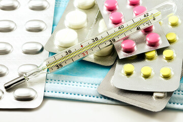 Blister packs with various medical pills on a medical mask. On top is a medical thermometer for temperature.