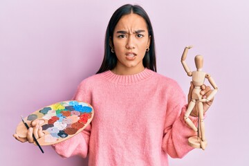 Young asian woman holding painter palette and art manikin clueless and confused expression. doubt...