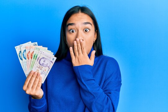 Young asian woman holding singapore dollars banknotes covering mouth with hand, shocked and afraid for mistake. surprised expression