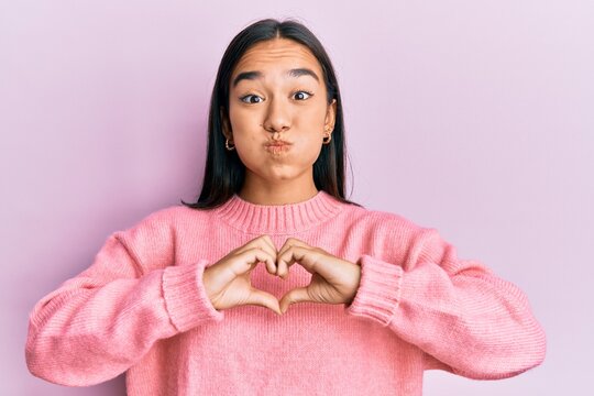 Young asian woman doing heart symbol with hands puffing cheeks with funny face. mouth inflated with air, catching air.