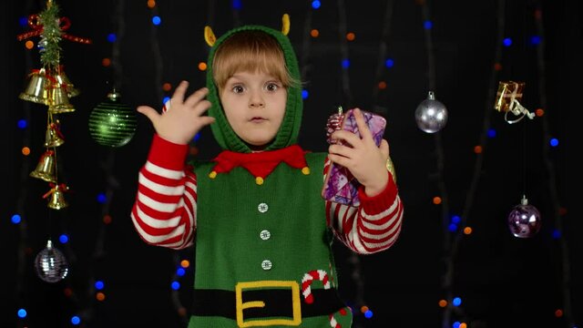 Little kid child girl in Christmas elf Santa Claus helper costume types something on mobile phone display enjoys browsing social media sends messages of congratulations, online gifts shooping sale