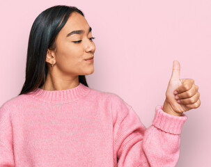 Young asian woman wearing casual winter sweater looking proud, smiling doing thumbs up gesture to the side