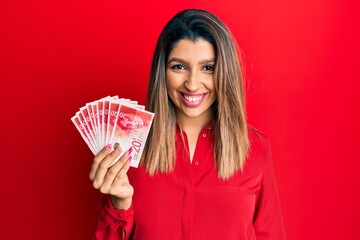 Beautiful brunette woman holding 20 israel shekels banknotes looking positive and happy standing...