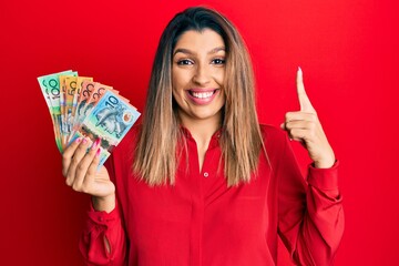 Beautiful brunette woman holding australian dollars smiling amazed and surprised and pointing up with fingers and raised arms.