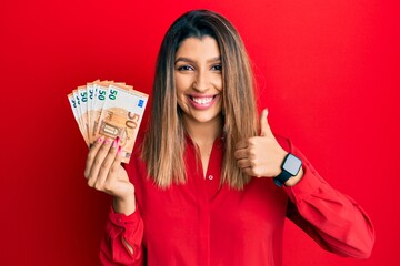 Beautiful brunette woman holding 50 euro banknotes smiling happy and positive, thumb up doing excellent and approval sign