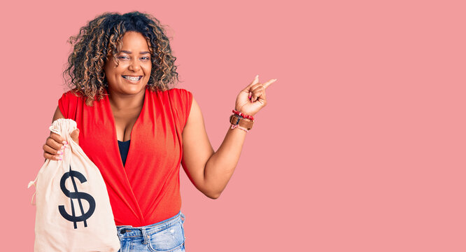 Young african american plus size woman holding money bag with dollar symbol smiling happy pointing with hand and finger to the side