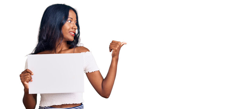 Young indian girl holding blank empty banner pointing thumb up to the side smiling happy with open mouth