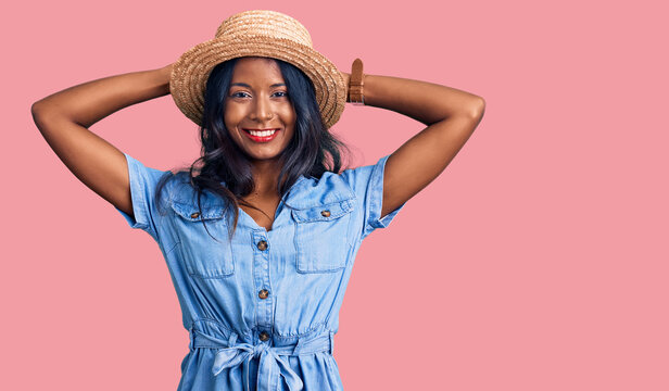Young indian girl wearing summer hat relaxing and stretching, arms and hands behind head and neck smiling happy
