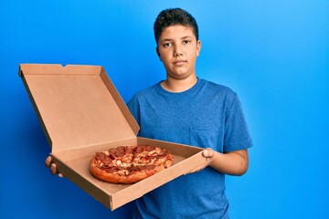 Teenager hispanic boy eating tasty pepperoni pizza relaxed with serious expression on face. simple and natural looking at the camera.