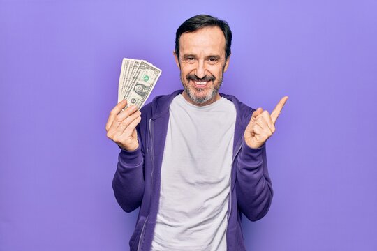 Middle age handsome man holding bunch of dollars banknotes over isolated purple background smiling happy pointing with hand and finger to the side