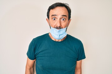 Middle age hispanic man wearing medical mask scared and amazed with open mouth for surprise, disbelief face