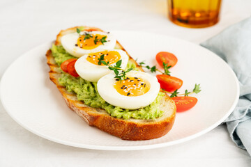 Avocado toast with toasted bread soft-boiled eggs with yellow yolk and tomatoes with herbs on white plate on light tablecloth