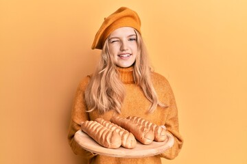 Beautiful young caucasian girl wearing french look with beret holding baguettes bread winking...