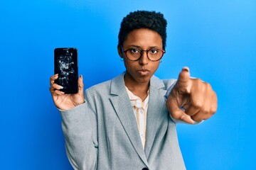 Young african american girl holding broken smartphone showing cracked screen pointing with finger to the camera and to you, confident gesture looking serious