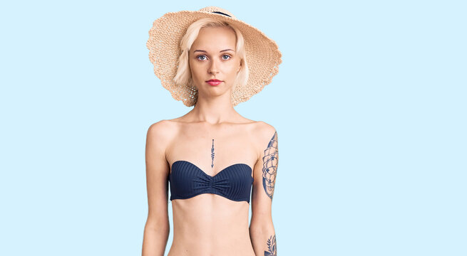 Young blonde woman with tattoo wearing bikini and summer hat relaxed with serious expression on face. simple and natural looking at the camera.