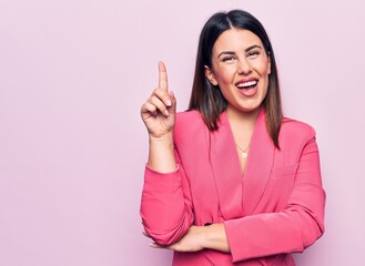 Young beautiful business woman wearing elegant jacket standing over pink background smiling with an idea or question pointing finger up with happy face, number one