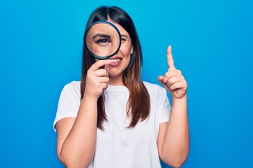 Young beautiful brunette woman using magnifying glass over isolated blue background smiling with an idea or question pointing finger with happy face, number one