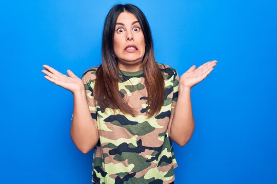Young beautiful brunette woman wearing camouflage military t-shirt over blue background clueless and confused with open arms, no idea and doubtful face.
