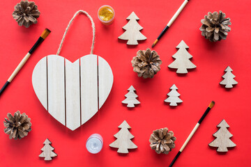 Christmas tree craft wooden eco friendly natural holiday concept. Heart shape wooden sign board frame hanging on a rope with little fir trees, pine cones, brushes and paints on red background.