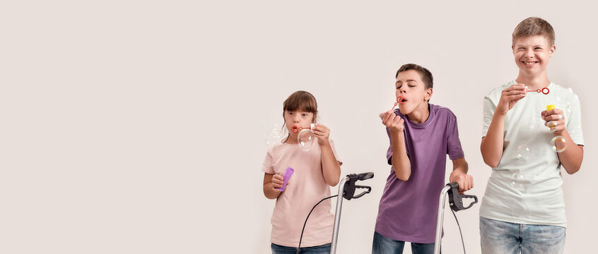 Three cheerful disabled children with Down syndrome and cerebral palsy smiling while blowing soap bubbles, standing together isolated over white background