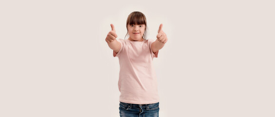 Portrait of disabled girl with Down syndrome smiling at camera, showing thumbs up with both hands while standing isolated over white background