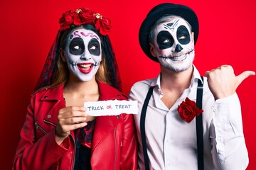 Couple wearing day of the dead costume holding trick or treat paper pointing thumb up to the side smiling happy with open mouth