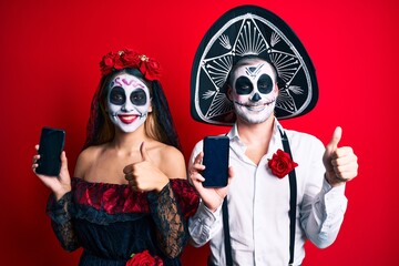 Couple wearing day of the dead costume holding smartphone showing screen smiling happy and positive, thumb up doing excellent and approval sign