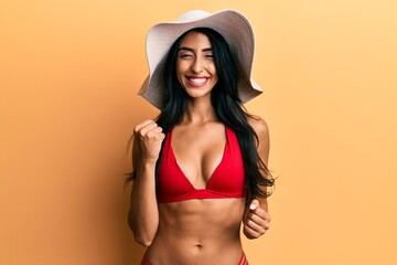 Beautiful hispanic woman wearing bikini and summer hat celebrating surprised and amazed for success with arms raised and eyes closed